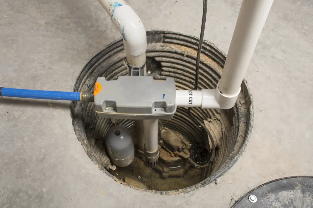 A sump pump system installed in a concrete basement floor, with PVC pipes and electrical components visible around and inside the pit.