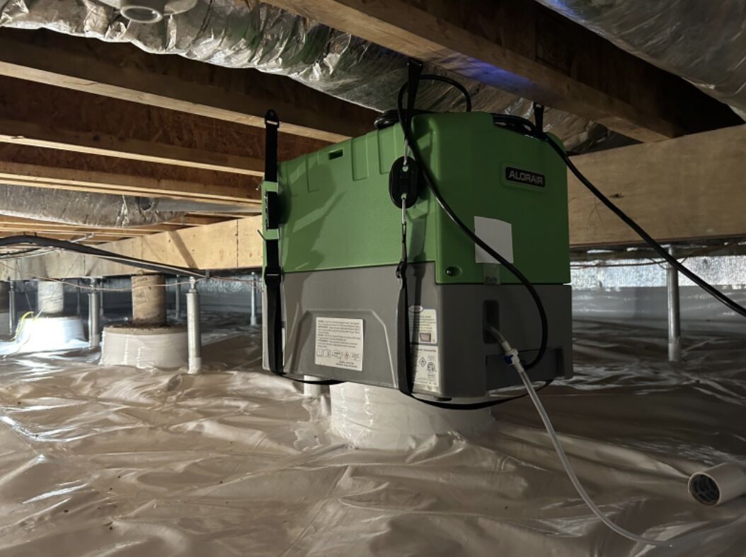 A green dehumidifier unit is installed under a house in a crawl space with plastic vapor barrier lining the floor and exposed wooden beams above.