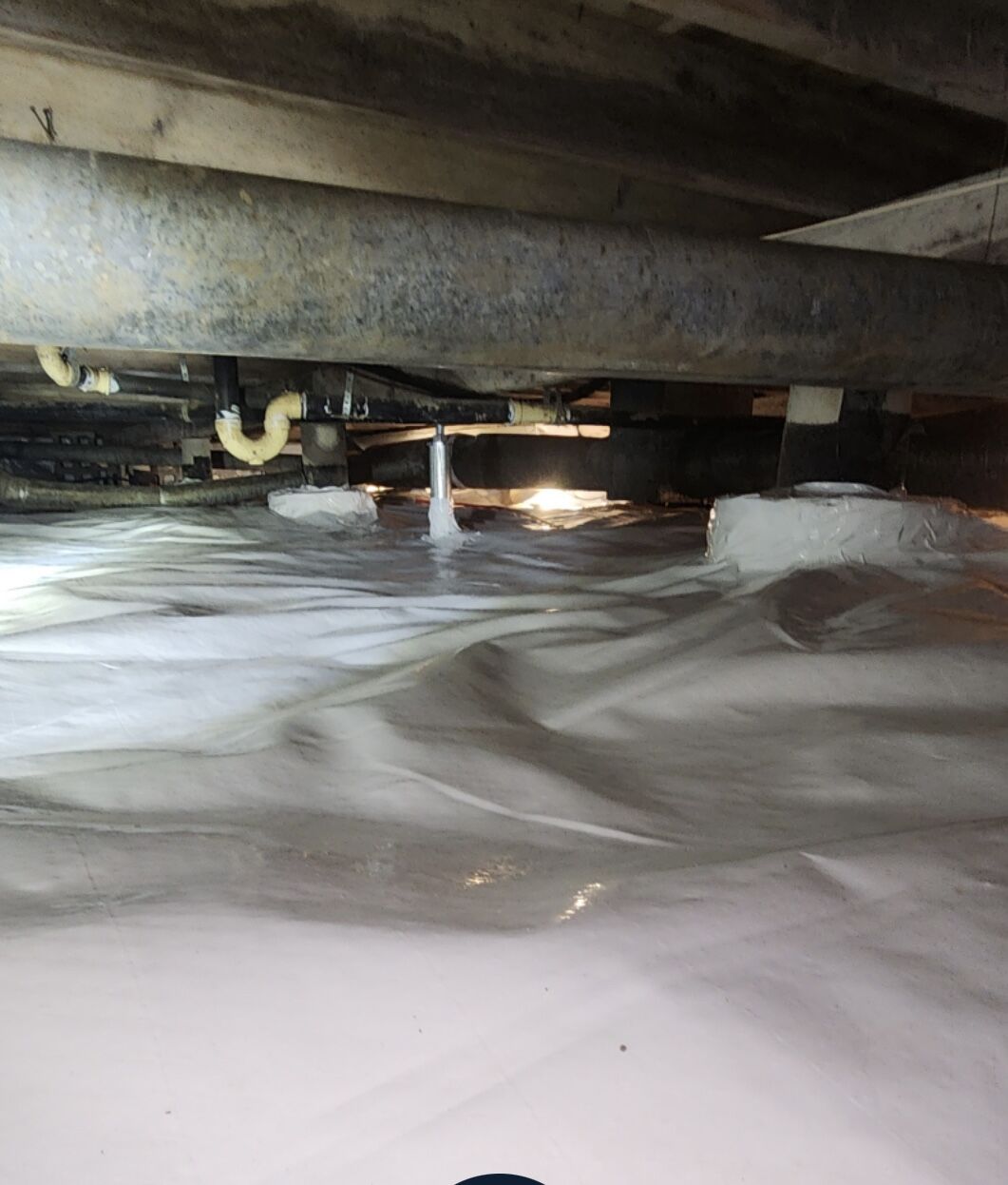 View of a crawl space with exposed pipes and beams above a white plastic vapor barrier covering the ground. Light is visible in the background.