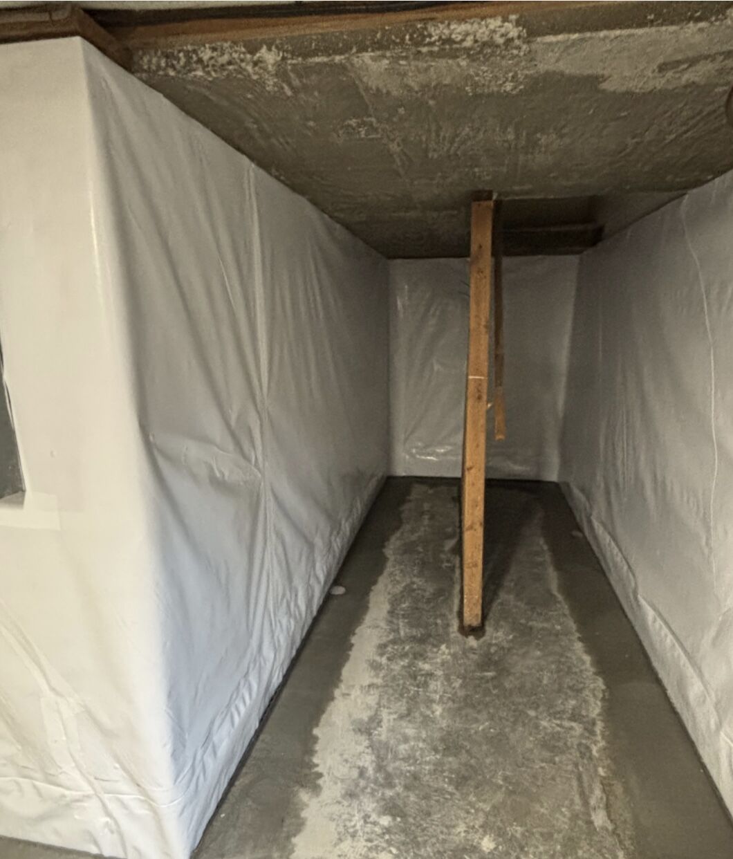 A narrow concrete room with plastic sheets covering the walls and ceiling, and a wooden beam supporting the ceiling in the center.