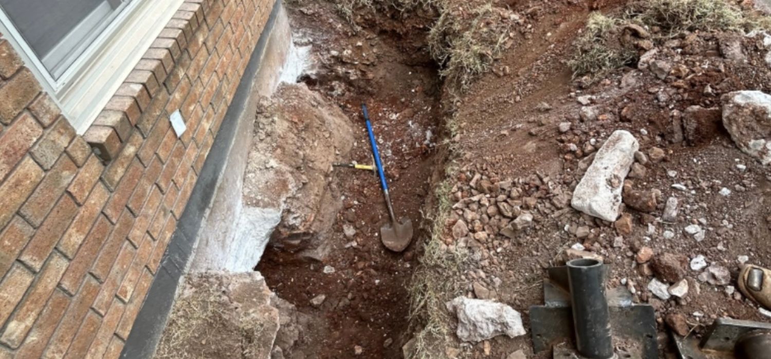 A trench with loose soil and rocks is dug next to a brick building&mdash;potential warning signs of foundation damage; a shovel and tape measure rest in the trench.