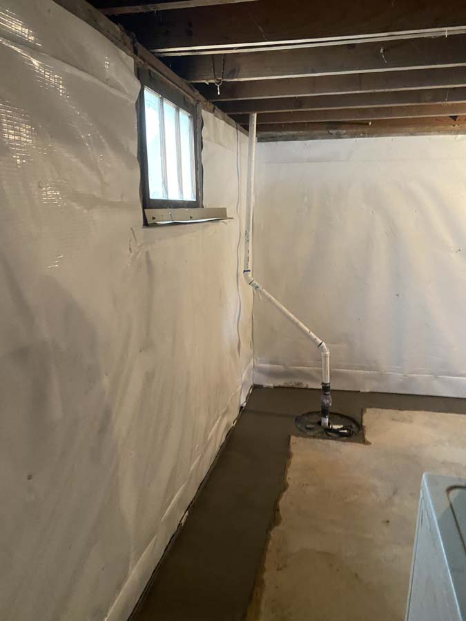 Basement corner with exposed ceiling beams, a small window, plastic-lined walls, cement floor, and a sump pump with connected piping.