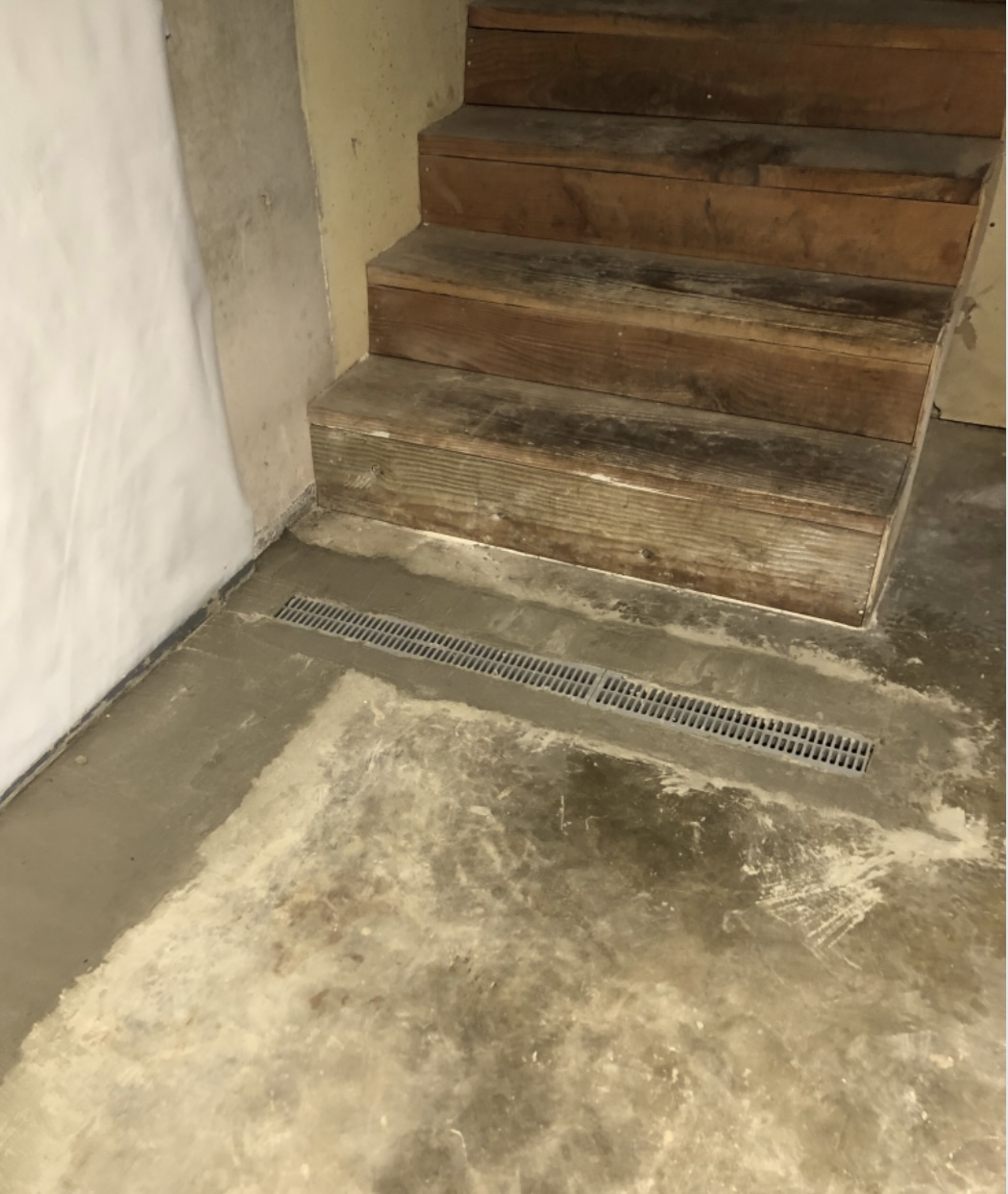 Wooden stairs lead up from a concrete basement floor with a metal floor drain grate nearby and unfinished walls in the background.