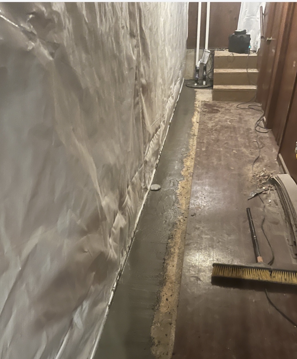 A basement area with a partially finished concrete strip along the wall, covered piping, tools, and a broom visible on the wooden floor.