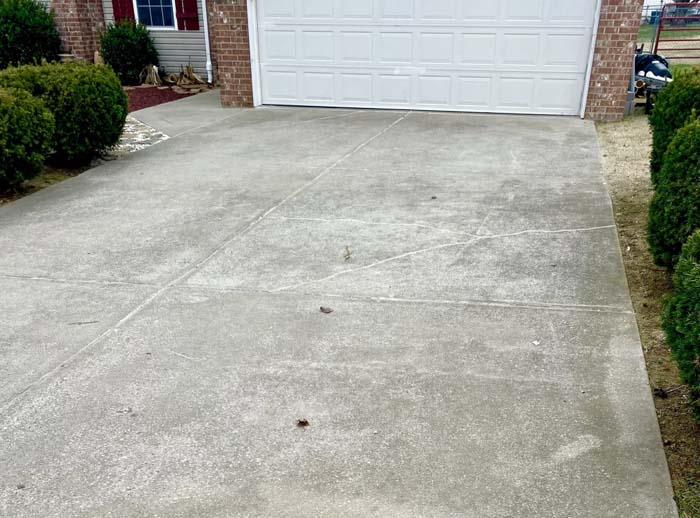 A concrete driveway with minor cracks and a few scattered leaves, leading up to a white garage door on a brick house. Shrubs line the sides of the driveway.