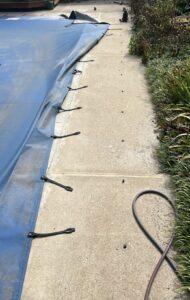 Blue pool cover partially installed on concrete pool deck, with black straps secured in anchor holes; power tool and cord visible nearby.