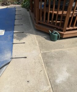 A green watering can sits on a concrete patio next to a wooden structure and a covered pool secured with black straps.