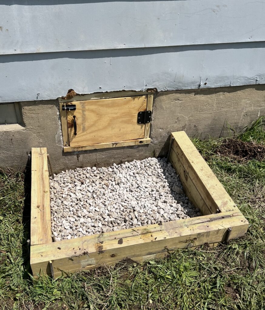 A small wooden frame filled with white gravel is placed against the foundation of a building, below a hinged wooden access door.