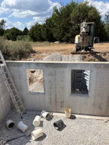 Concrete foundation wall with two window openings, buckets and a ladder inside, and an excavator positioned on gravel outside under a partly cloudy sky.