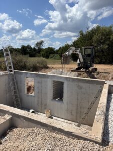 A small excavator is parked near a concrete foundation under construction, with ladders propped against the structure and gravel piled nearby.