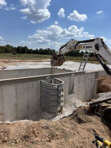 An excavator lowers a prefabricated basement window well into place against a concrete foundation wall at a construction site under a partly cloudy sky.