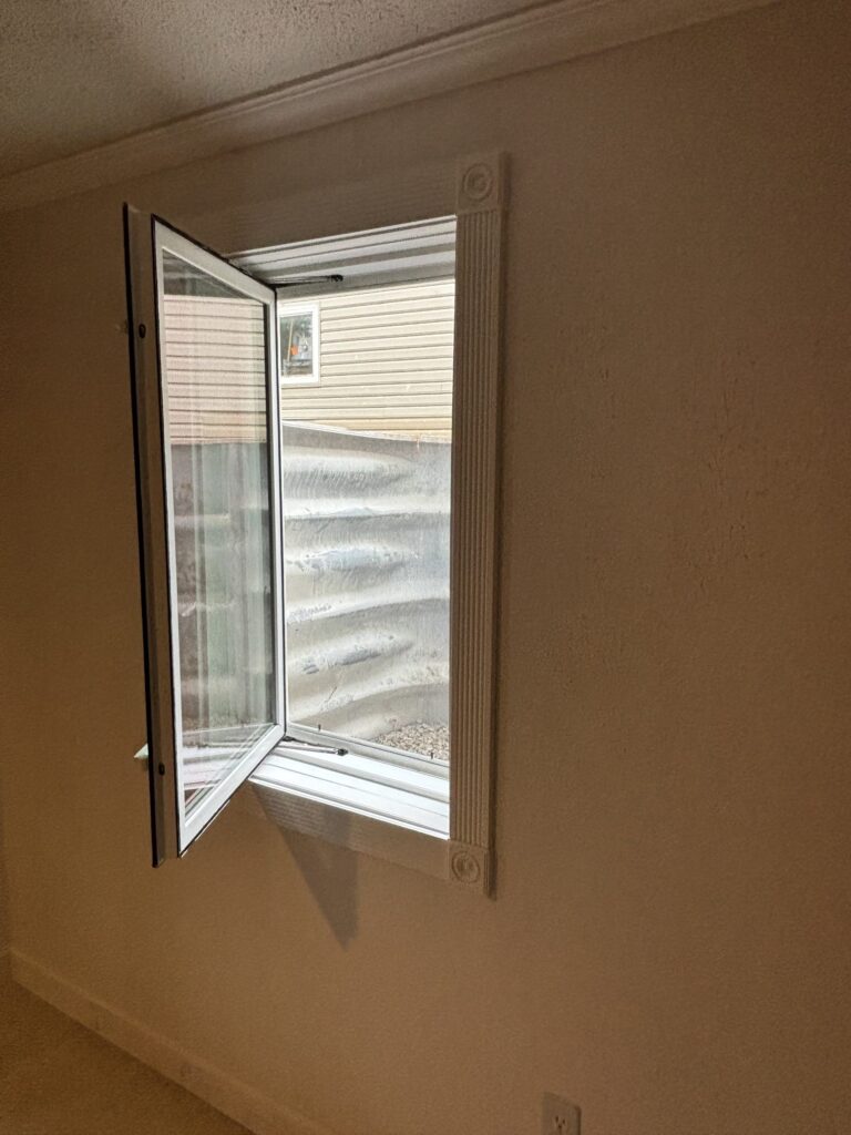Open casement window in a beige room, showing an outdoor window well and neighboring building.