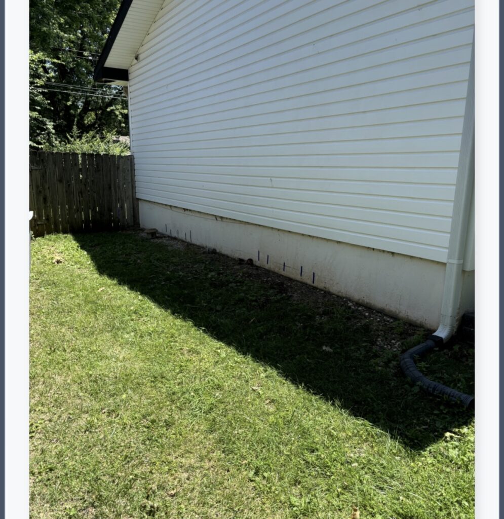 Side of a white building with a sloped roof, grassy lawn, small fence, and a drainage pipe.