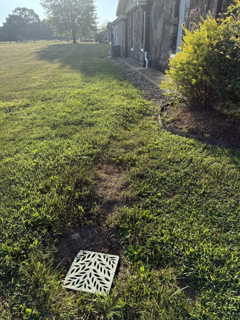 A decorative metal drain cover is set in a grassy yard near a house with stone siding and shrubbery on a sunny day.