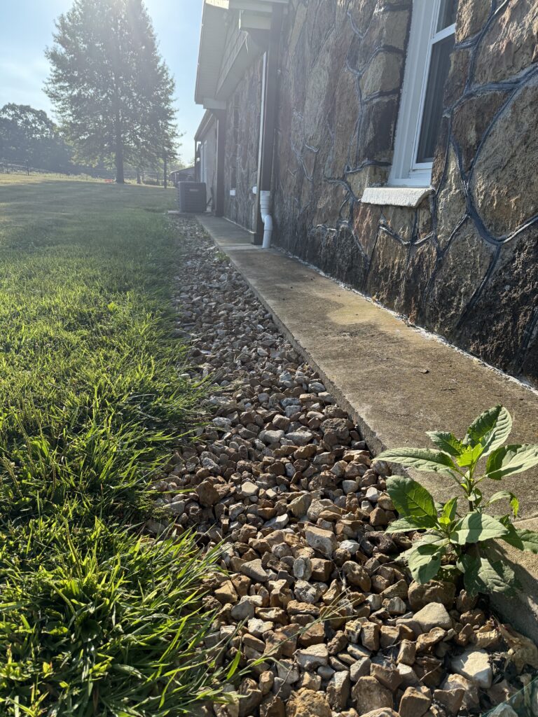 A concrete walkway runs alongside a stone house, bordered by a strip of rocks and a patch of grass, with a small weed growing near the edge.