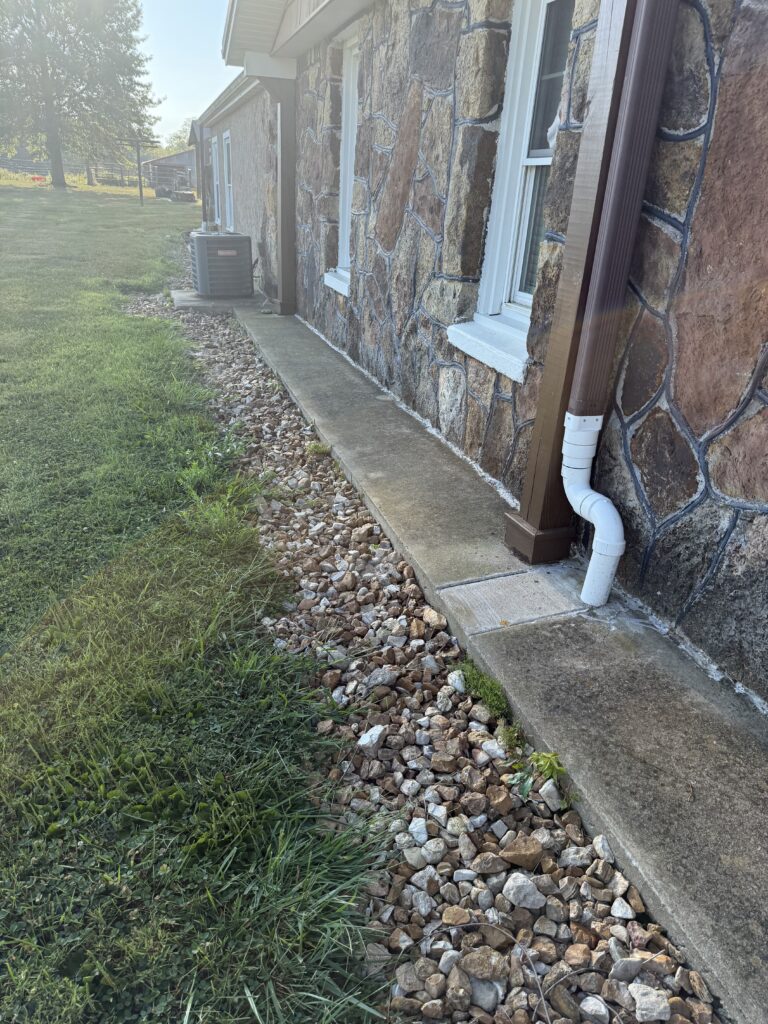 A stone walkway runs alongside a house with stone siding, bordered by rocks and grass, with a downspout pipe and an outdoor air conditioning unit visible.