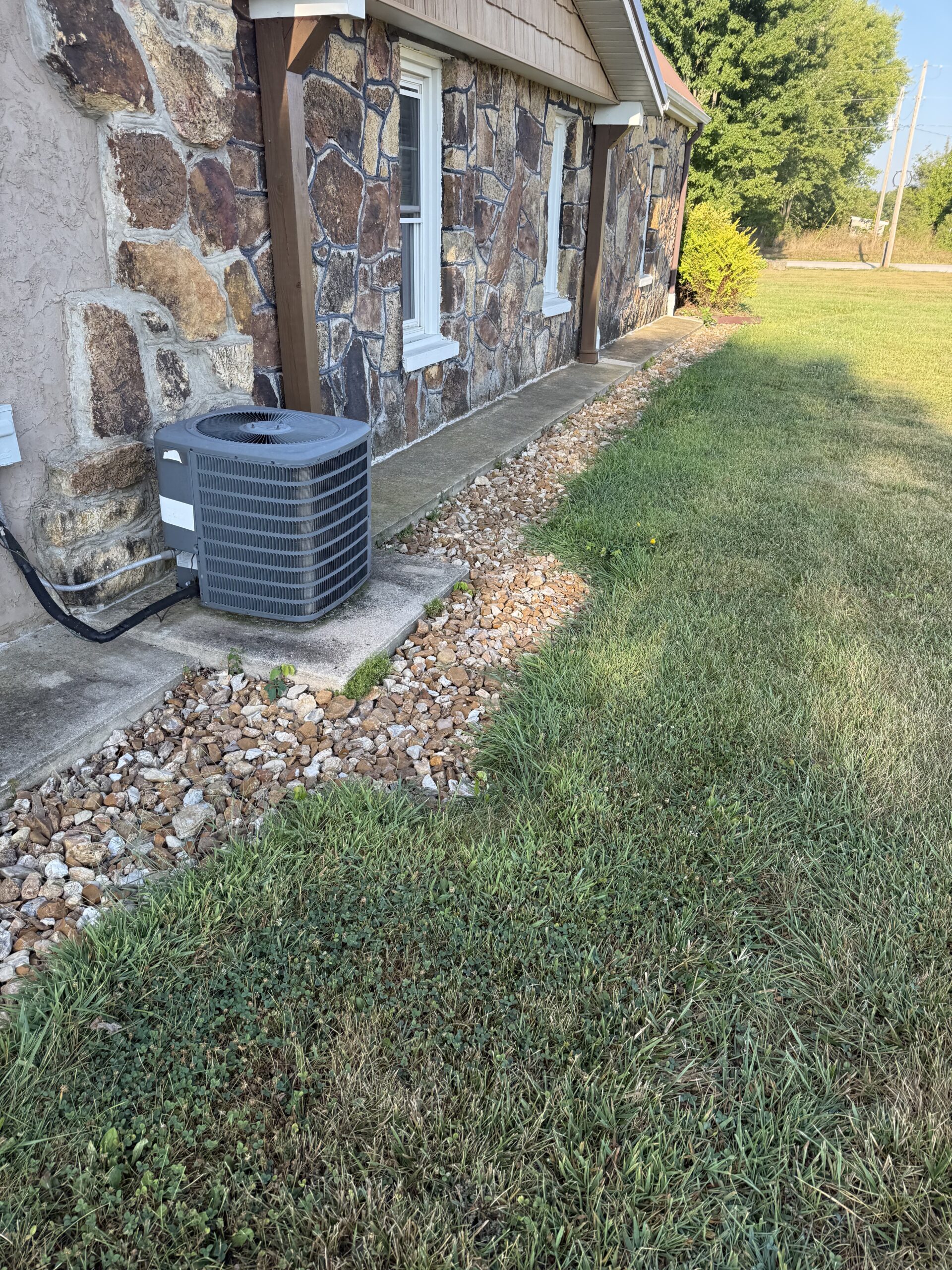 An outdoor air conditioning unit sits on a concrete pad beside a stone house, with a border of rocks separating the lawn from the building.