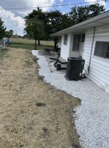 White gravel borders the foundation of a white house with an air conditioning unit outside; the adjacent yard has patches of dry grass and dirt.