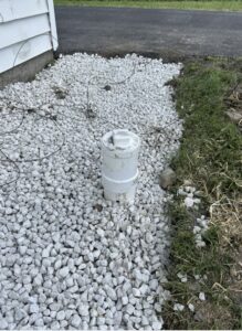 A white PVC cleanout pipe extends from gravel near a house, next to a patch of grass and a paved driveway.