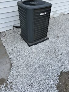 A central air conditioning unit sits on a bed of white gravel beside a white-sided building.