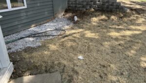 A yard with straw-covered ground, a patch of white gravel near a house, a garden hose, a small white drainpipe, and a retaining wall in the background.