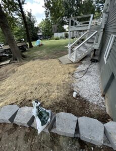 A backyard with newly seeded soil covered in straw, concrete pavers in the foreground, a bag of lawn care product, and a staircase leading to a raised deck on the right.
