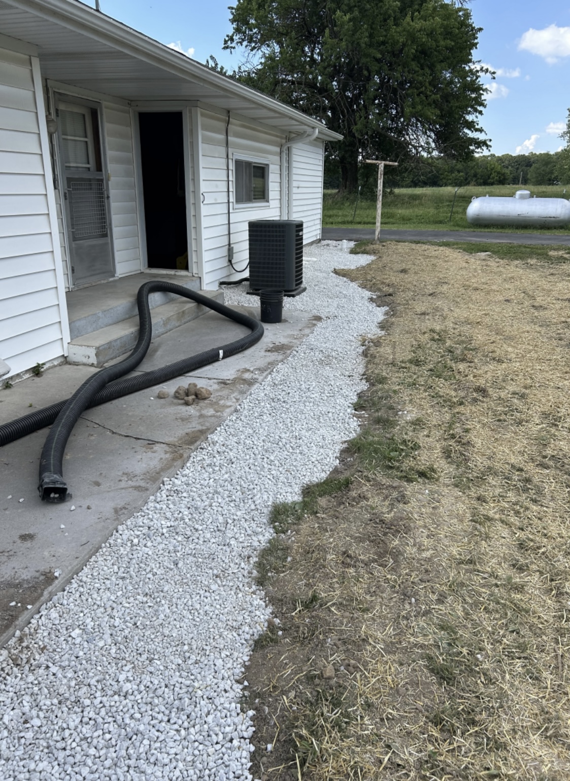 A gravel path runs alongside a white house with a black hose and air conditioning unit near the door; a propane tank is visible in the yard.
