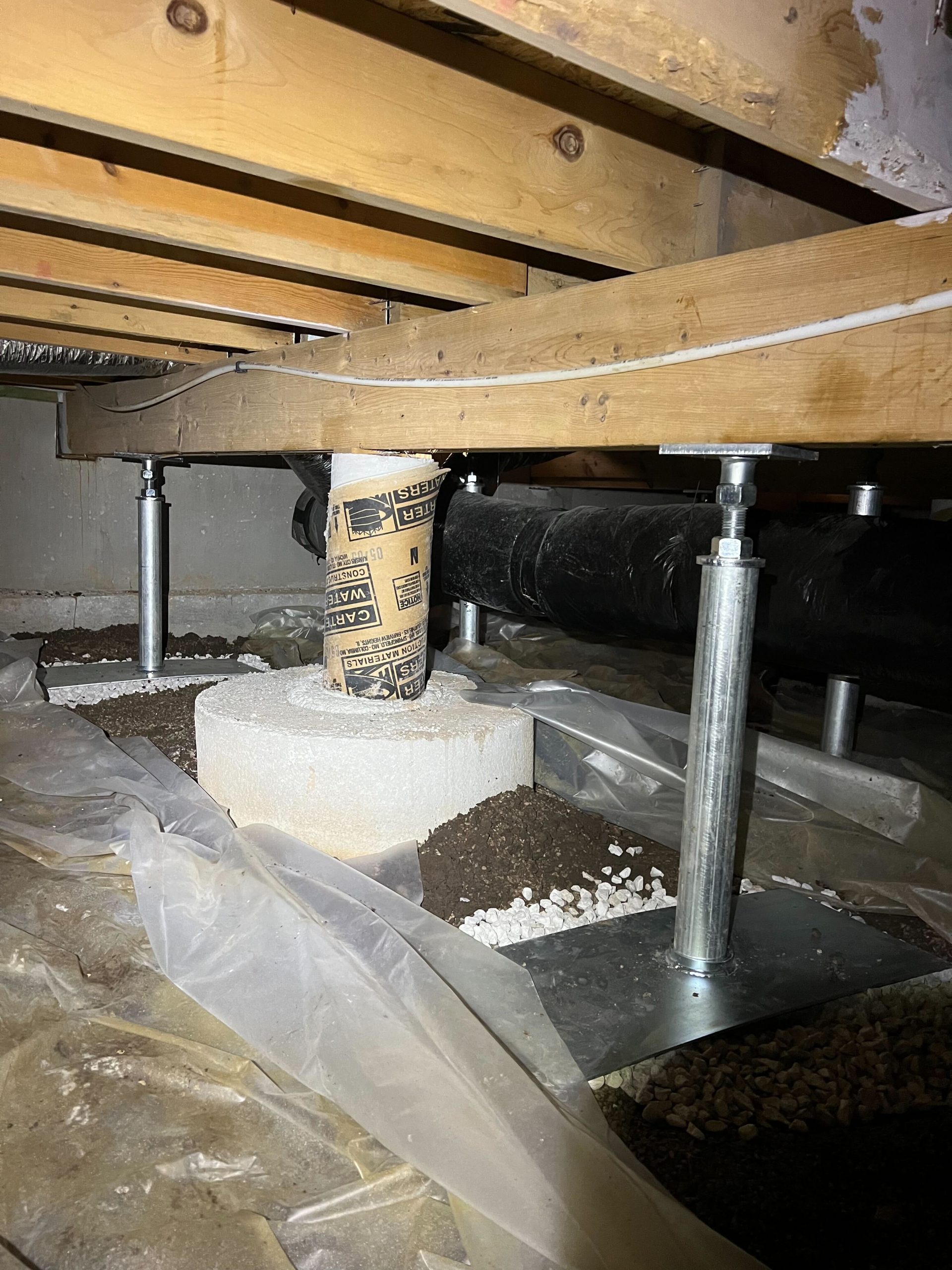 A crawl space with wooden beams, metal supports, and a concrete pillar wrapped in cardboard. Plastic sheeting covers the ground, with small gravel piles spread across the area.