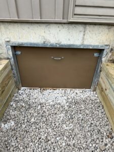 Metal hatch door set in a concrete foundation, surrounded by gravel and wooden borders, with siding above.