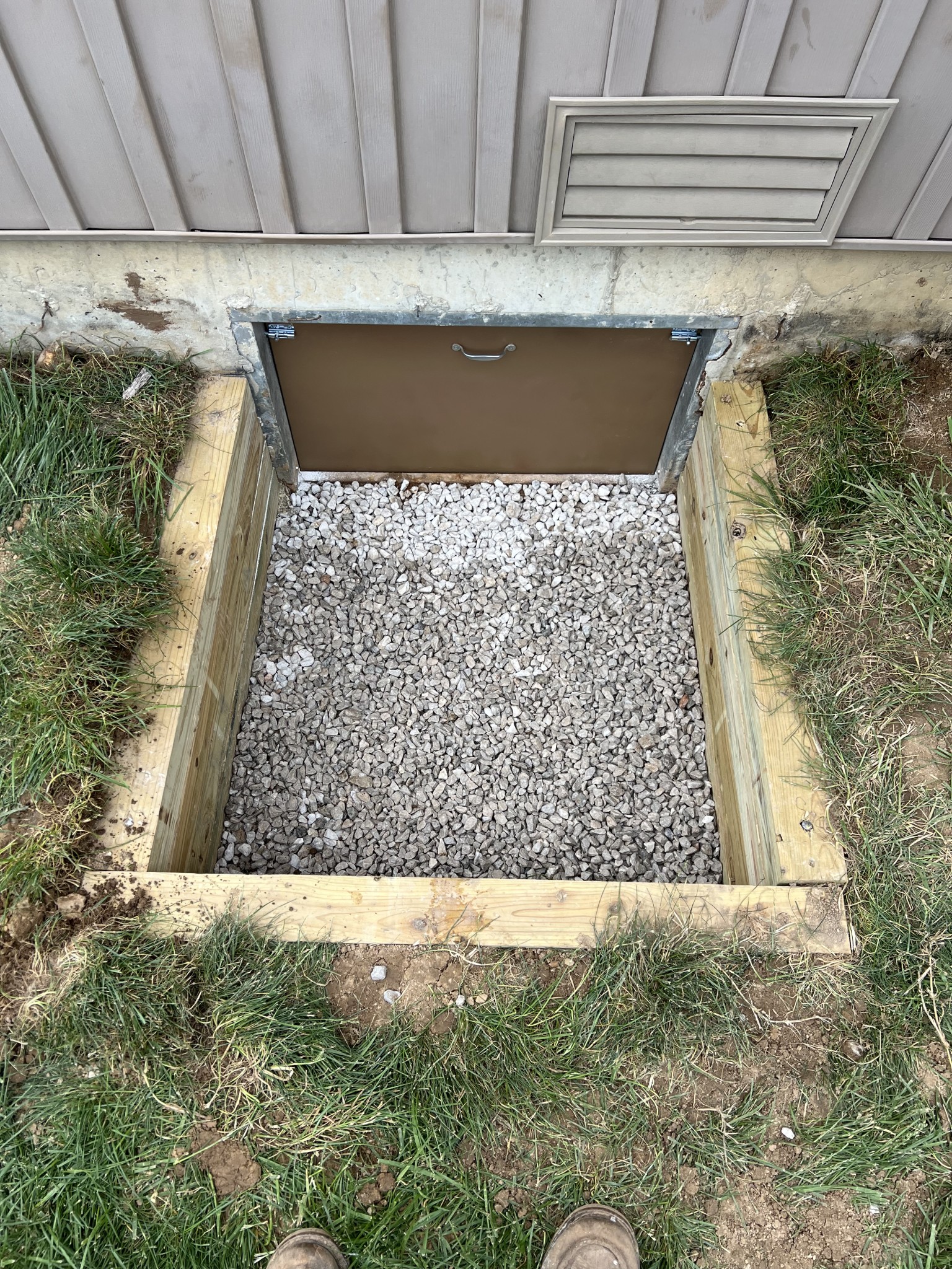 A basement egress window with a metal cover, surrounded by a wooden frame and filled with gravel, set in a grassy area.