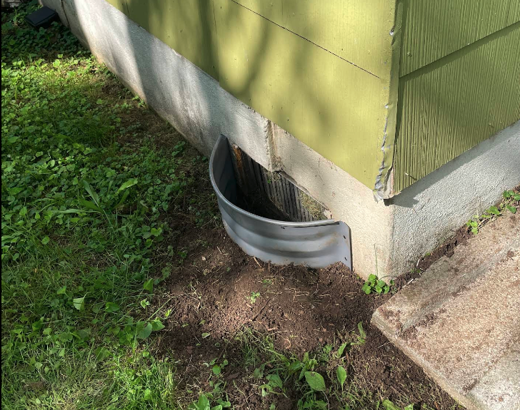 A metal window well is attached to a basement window on a building with green siding, surrounded by grass and soil.