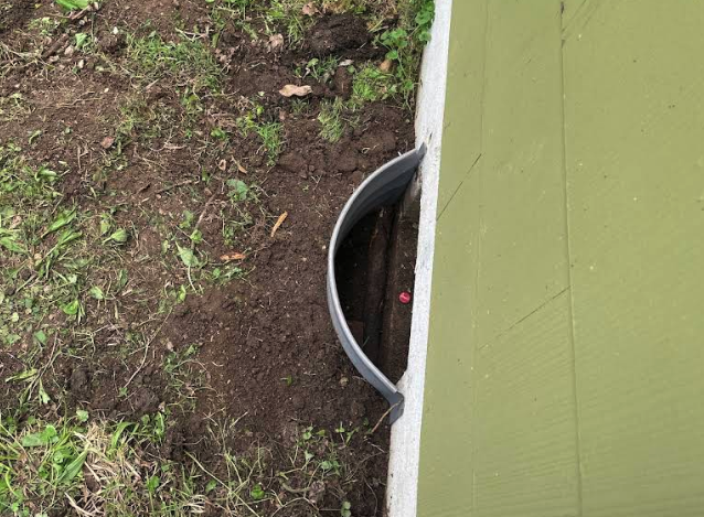 A partially buried pipe near the green exterior wall of a building, surrounded by soil and grass.
