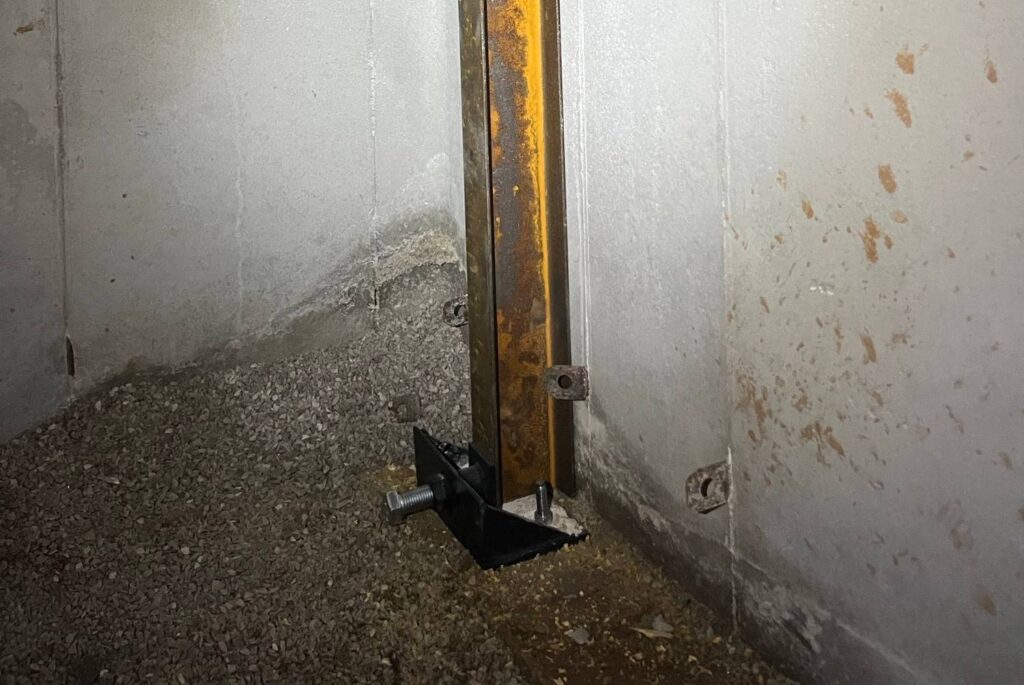 A steel support beam, functioning as one of the wall stabilizers, is anchored to a concrete basement wall and floor, showing signs of rust and adjacent wall cracks.