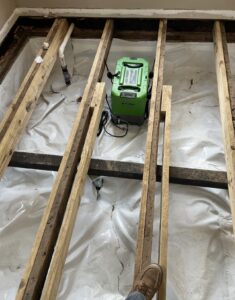 A green dehumidifier sits on plastic sheeting between exposed wooden floor joists in a room under renovation.