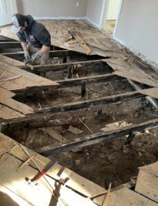 A worker removes damaged wooden floorboards, revealing the subfloor and joists in a room under renovation. Tools and debris are scattered around the work area.