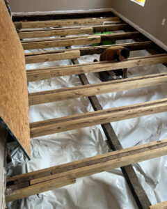 A person installs wooden floor joists over a plastic moisture barrier in a room with exposed subfloor.