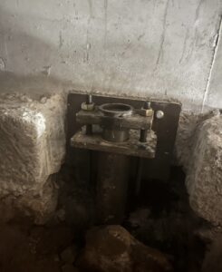 A close-up view of a steel foundation pier anchored to a concrete wall with bolts, partially surrounded by exposed soil and rough concrete edges.