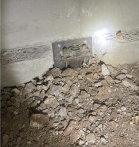 A metal electrical outlet partially embedded in a concrete wall, surrounded by uneven gravel and rocks, illuminated by a flashlight.