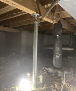 A steel adjustable support column installed under wooden floor joists in a crawl space, with a light source on the ground and visible dirt and debris.