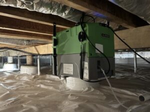 A green dehumidifier unit is installed under a house in a crawl space with plastic vapor barrier lining the floor and exposed wooden beams above.