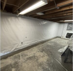 A basement with exposed ceiling beams, concrete floor, white wall covering, fluorescent lighting, and a partially covered fireplace in the corner.