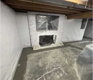 A basement with exposed ceiling beams, a white-painted brick fireplace, plastic coverings, and fresh concrete along the perimeter of the floor.