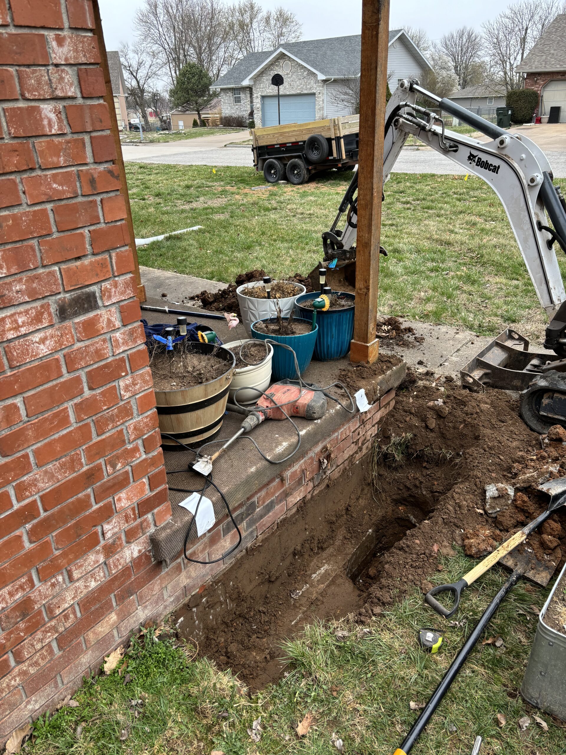 A small excavator digs near a brick house with tools, buckets, and a garden hose on the porch; a hole is visible next to the foundation.