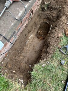 A trench dug alongside a brick wall exposes a white pipe underground; a shovel, tape measure, and power tool are nearby on the grass and pavement.