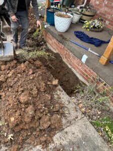 A person stands next to a freshly dug trench by a brick porch, with soil piled nearby and potted plants on the porch.