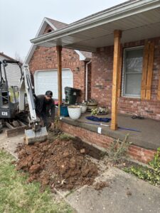 A man operates a small excavator, digging up soil near the porch of a brick house with various items and tools placed on the porch.