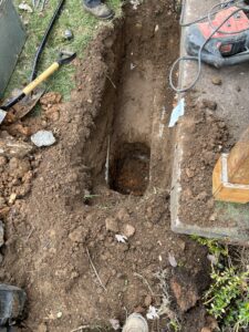 A freshly dug trench with a deeper hole at one end, surrounded by dirt, tools, and a concrete slab, next to a wooden post and some grass.