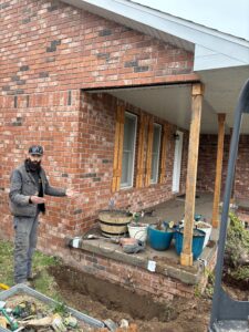 A man stands by a brick house with exposed foundation and construction tools, indicating ongoing work on the front porch area.