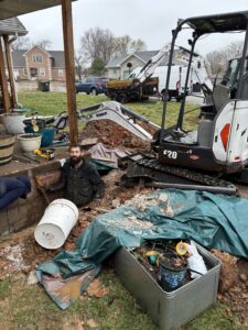 A man works in a muddy yard near a partially built brick wall, surrounded by construction tools, buckets, tarps, debris, and excavators.