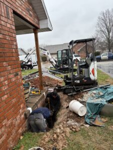 Two workers dig near the foundation of a brick house while a mini excavator and construction equipment are positioned nearby on a cloudy day.